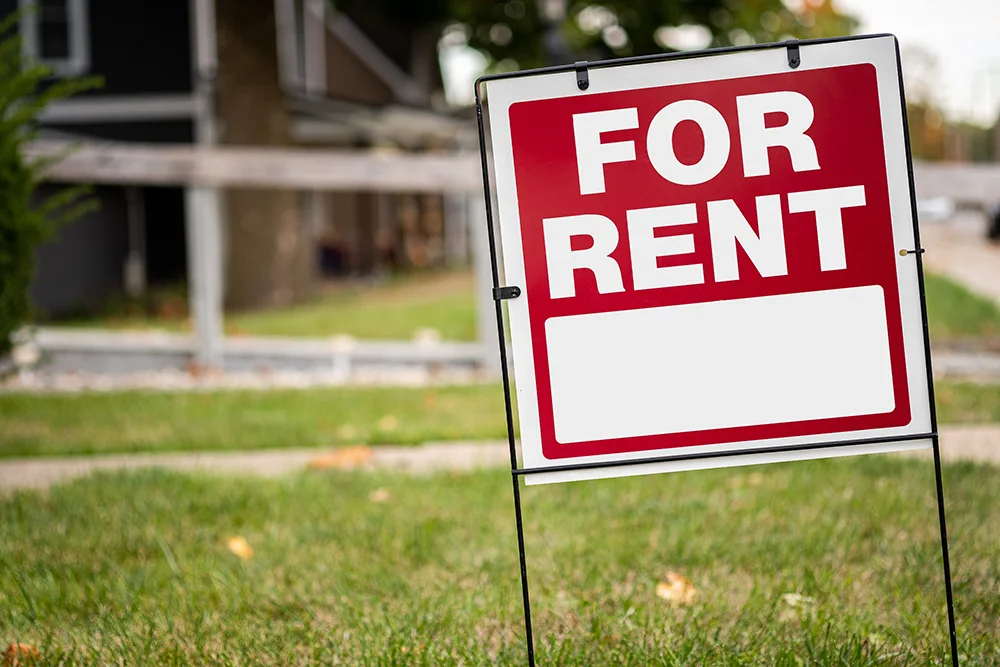 A red "FOR RENT" sign pictured on a lawn with homes in the background.