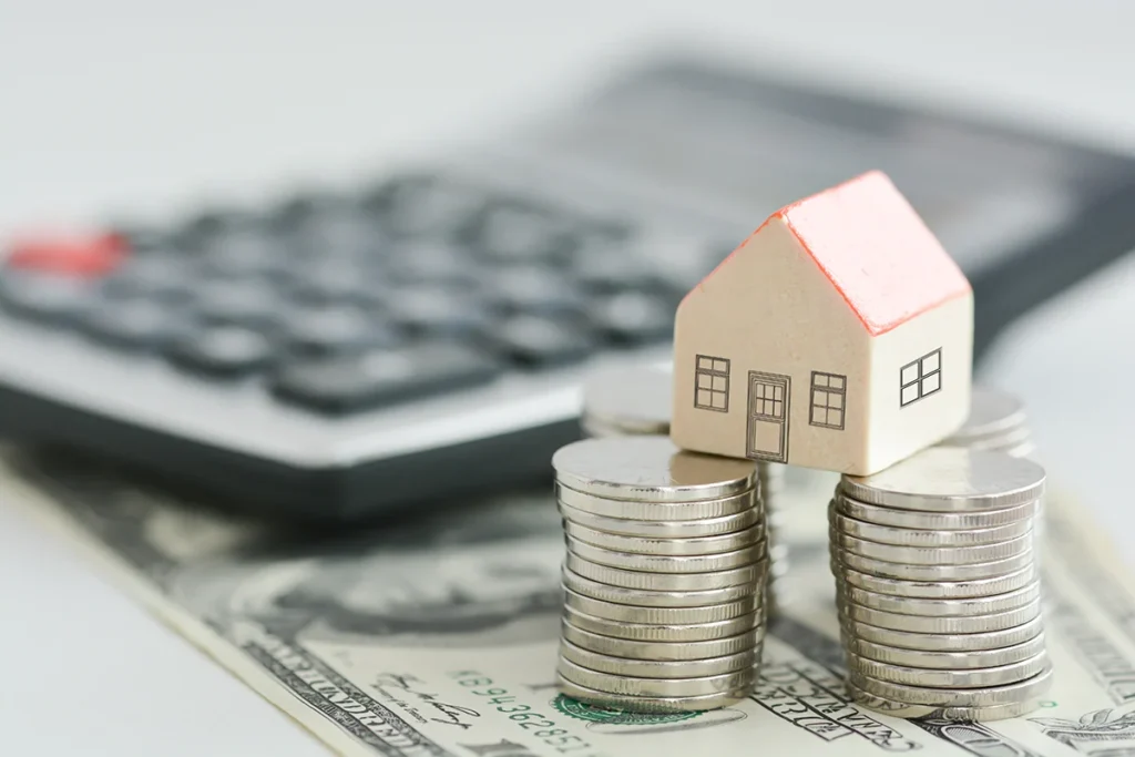 Coins with a toy house perched on top in the foreground, with a calculator in the background.